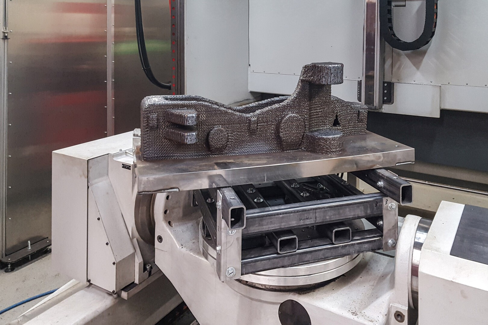 A large, 3D-printed metal industrial part sits on a rotating platform in a manufacturing machine, surrounded by mechanical components and metallic surfaces.