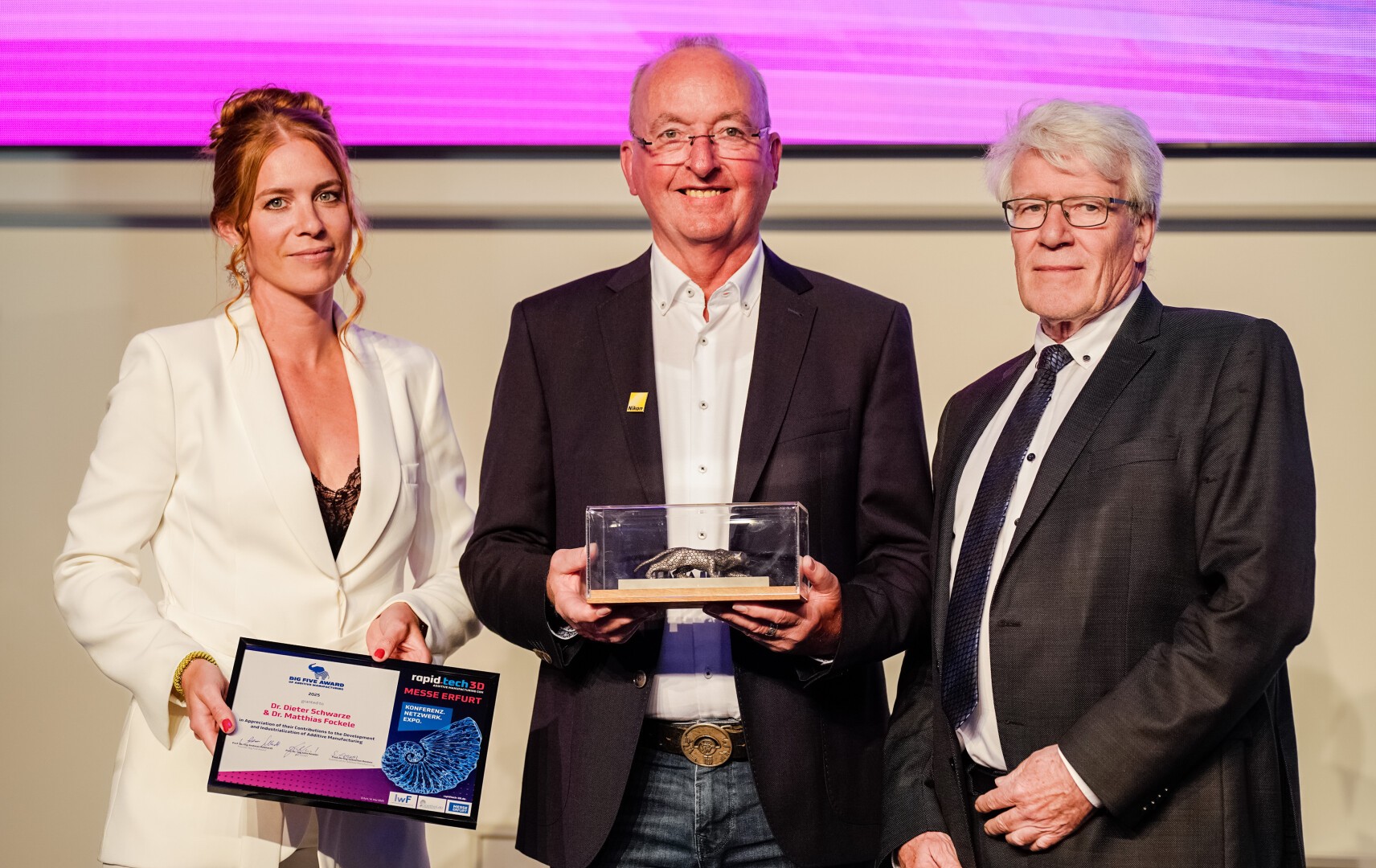 Three people in formal attire stand together at an award ceremony. The woman on the left holds a certificate, the man in the center holds a plaque in a case, and the man on the right stands with his hands together.