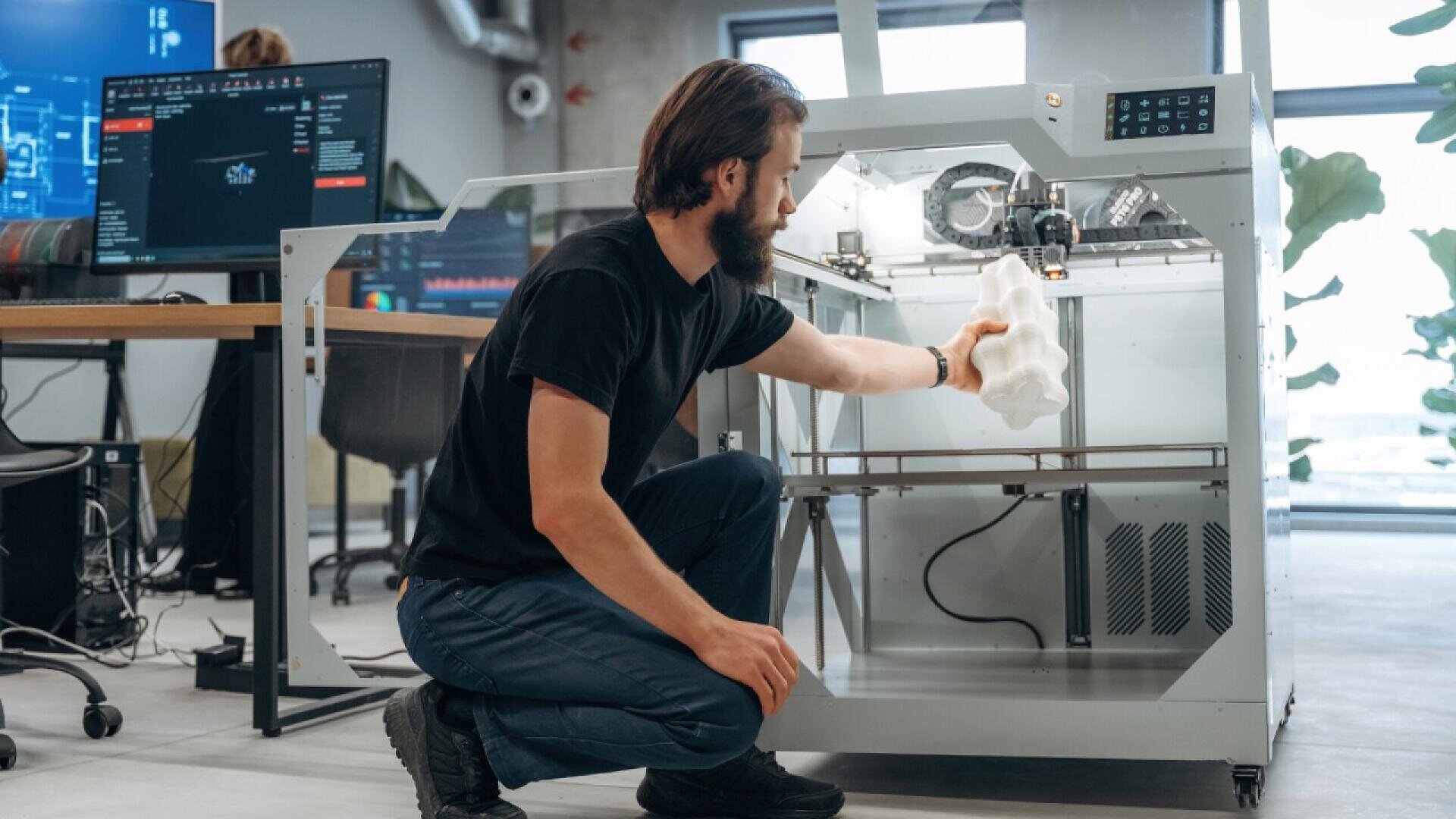 A man kneels beside a large 3D printer, removing a white geometric object from inside. Computer monitors and office equipment are visible in the background of a modern workspace.
