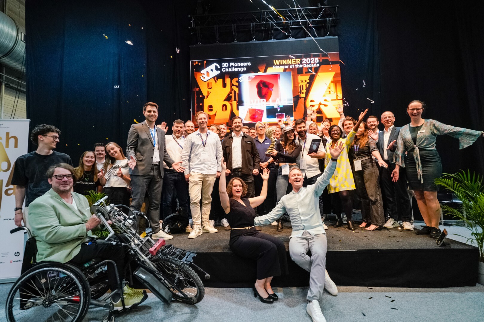 A large group of people smiling and celebrating on a stage, with confetti falling. Some hold trophies and a flag. A man in a wheelchair with a custom bike is at the front. A screen behind them displays Winner 2025 and event branding.