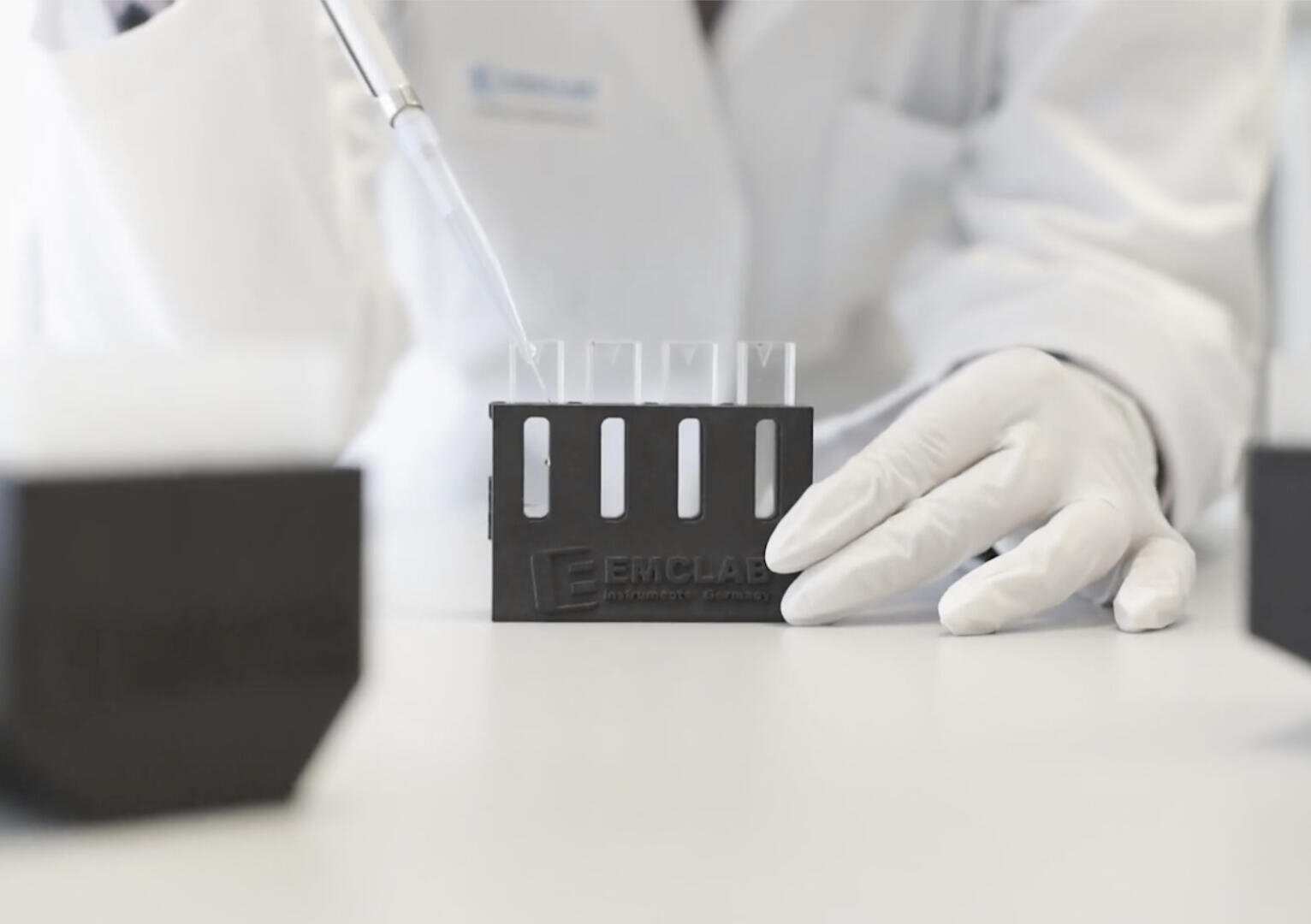 Eine Person in einem weißen Laborkittel und Handschuhen gibt mit einer Pipette Flüssigkeit in Reagenzgläser, die in einem schwarzen Gestell mit der Aufschrift „EMCLAE“ auf einer Laborwerkbank stehen.