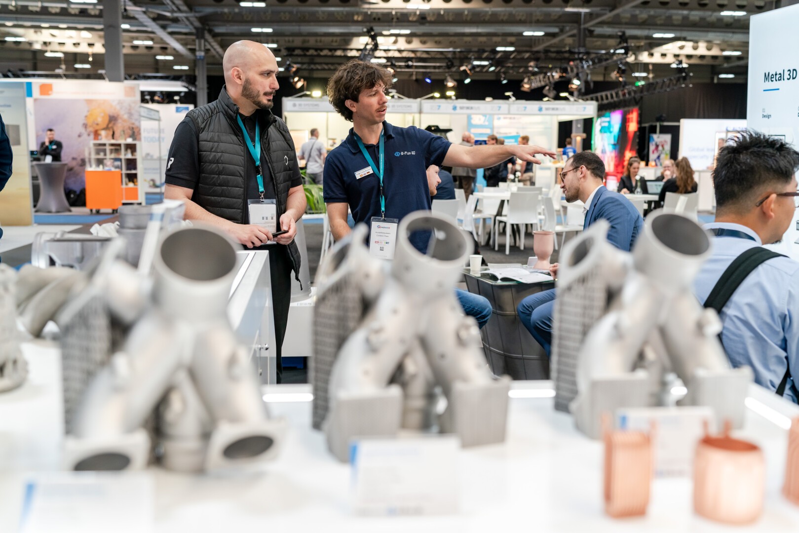 Two men stand near a table displaying metal 3D-printed parts at a technology expo. One man gestures while talking to the other; attendees and booths are visible in the busy background.