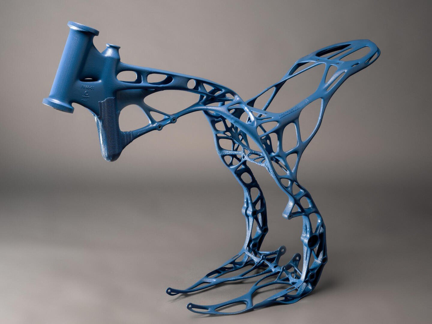A blue, futuristic bicycle frame with an organic, skeletal design structure stands upright against a neutral background. The frame features intricate curves and cutouts, resembling a blend of art and engineering.