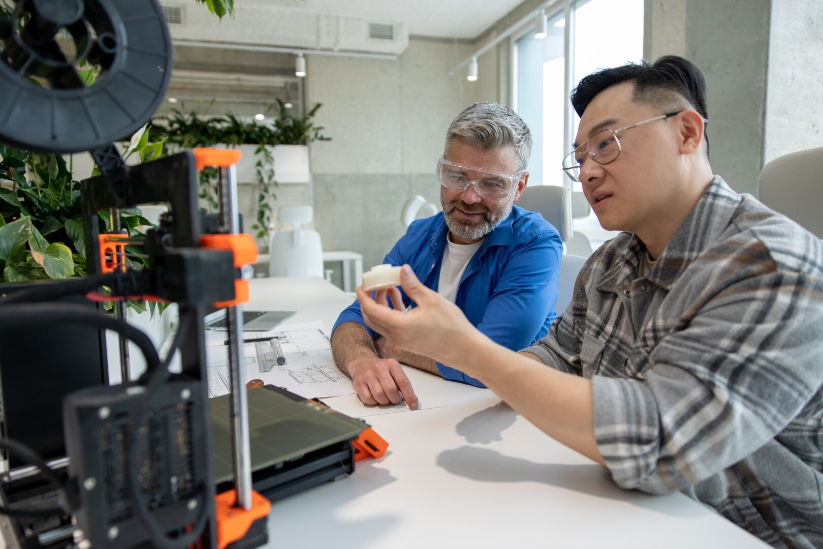Two men sit at a table examining a small 3D-printed object; one holds the object while the other looks on. A 3D printer, documents, and green plants are visible in the bright, modern workspace.