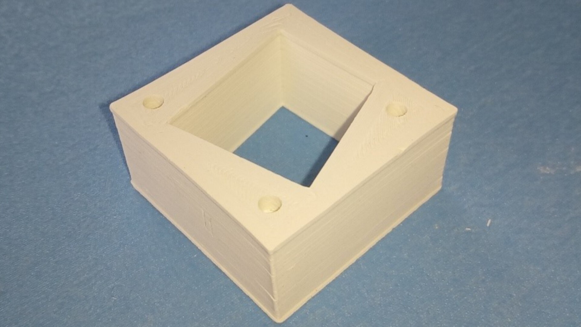 A 3D-printed, square hollow object with a square hole in the center sits on a blue surface. The object has four small circular marks, one near each corner on the top face.