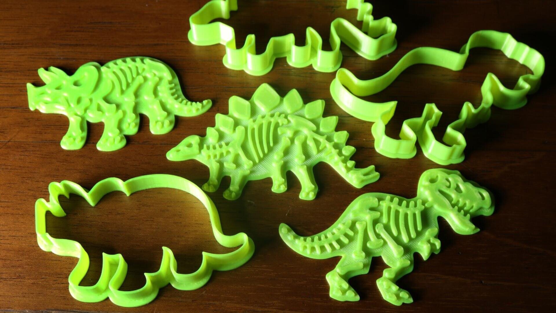Five green dinosaur-shaped cookie cutters, two showing detailed skeleton designs and three with simple outlines, are arranged on a wooden surface.