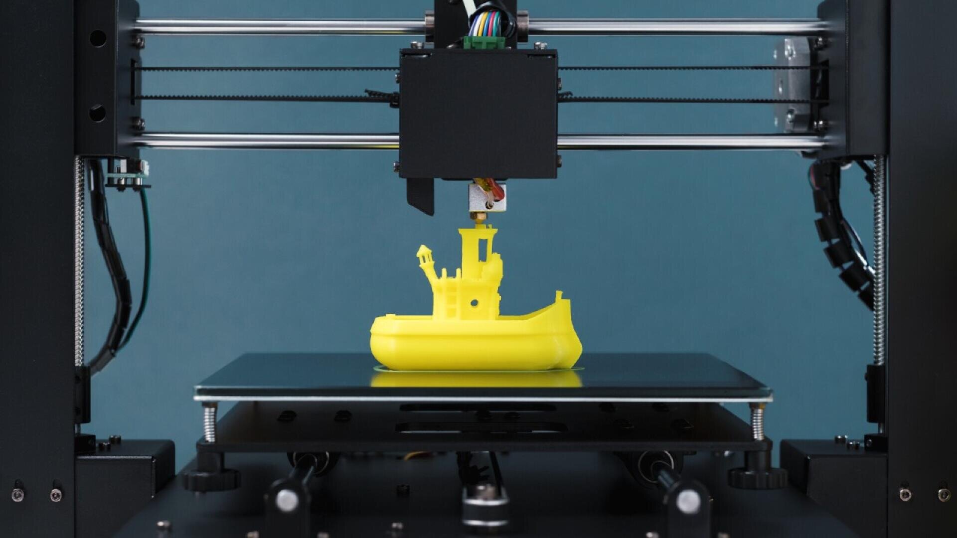 A 3D printer is creating a bright yellow boat model. The boat is partially finished and sits on the printers build plate against a plain blue background.