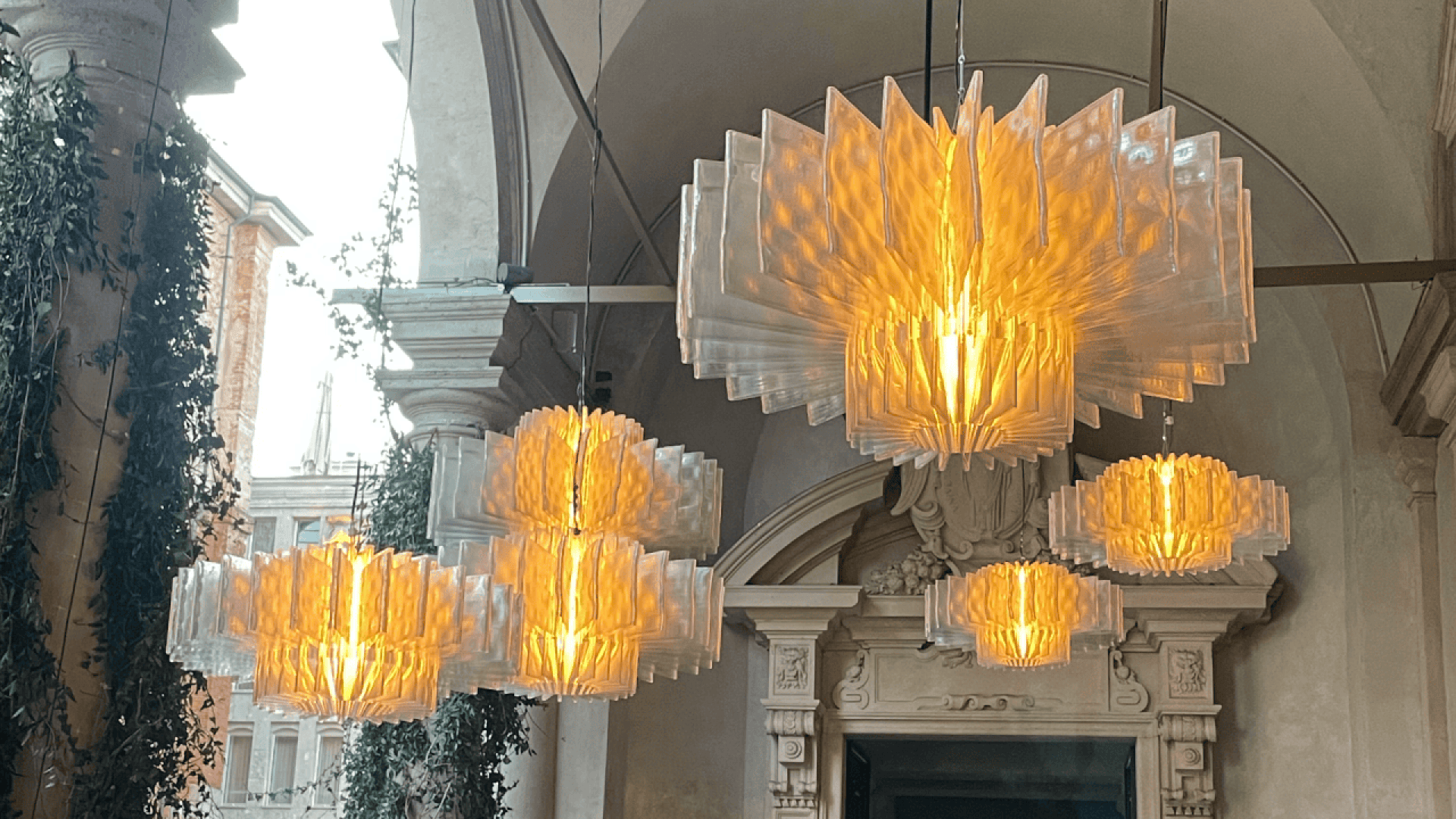 Large, modern chandeliers with translucent, geometric panels glow warmly under an arched ceiling adorned with classical architectural details and climbing greenery.