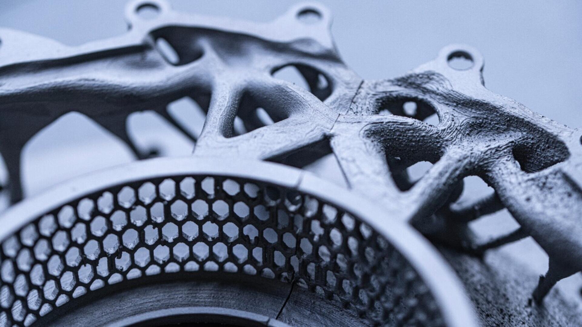 Close-up of a metallic 3D-printed component, featuring a honeycomb pattern and intricate, organic lattice structures with a smooth and textured finish. The image highlights fine details and advanced engineering design.