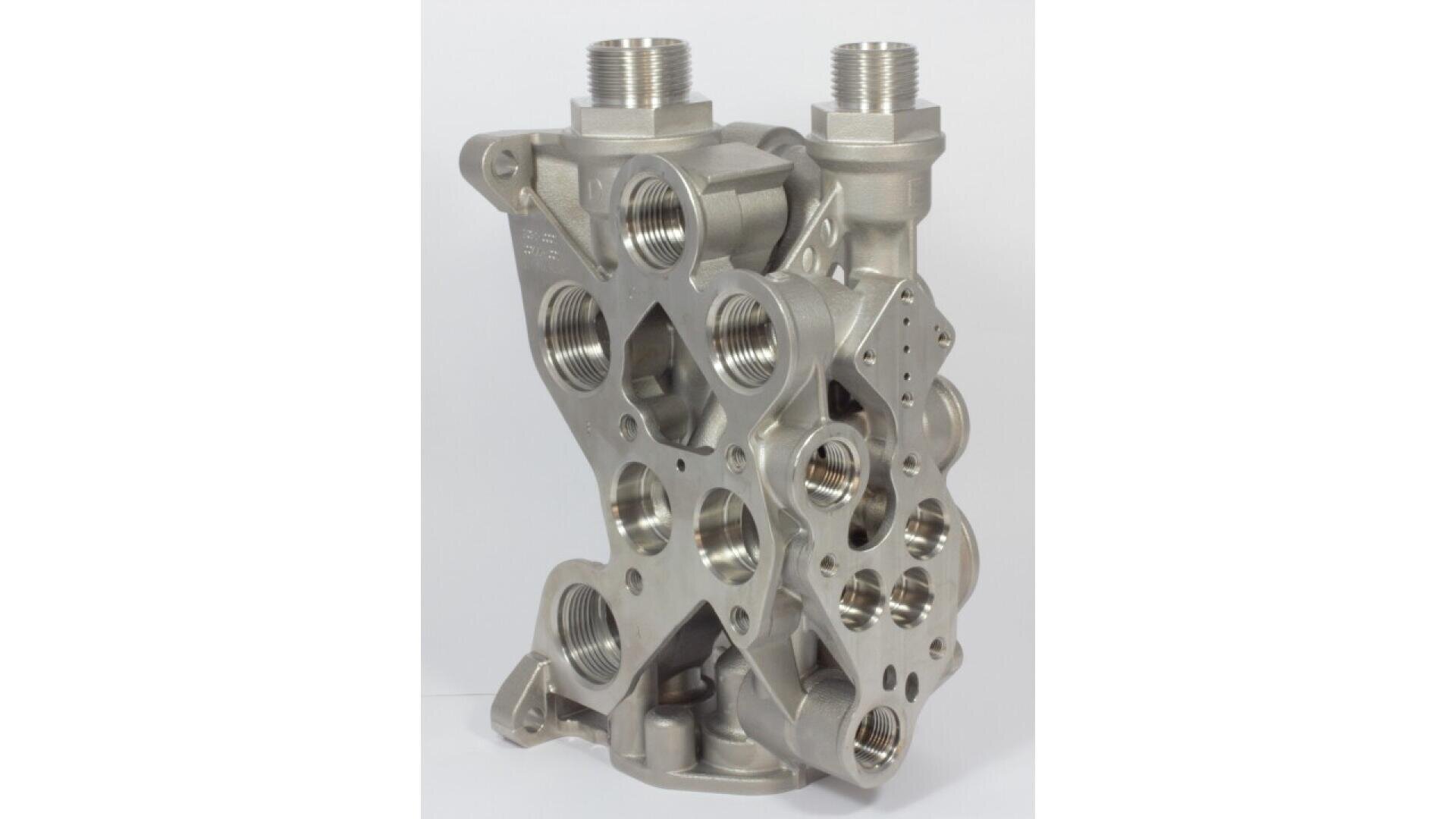 A metallic, industrial valve block with multiple hexagonal and circular holes and threaded pipe connections, standing vertically against a plain white background.