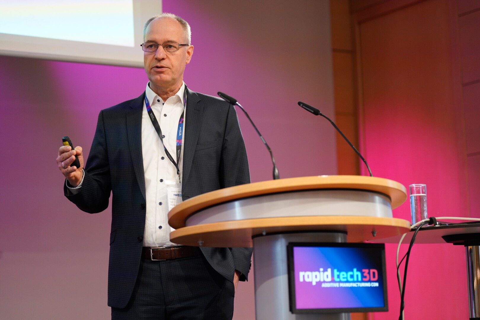 Ein Mann im Anzug steht an einem Podium und hält einen Vortrag bei der Rapid Tech 3D-Veranstaltung. Er hält eine Fernbedienung in der Hand. Zwei Mikrofone stehen auf dem Podium, neben ihm steht ein Glas Wasser. Der Hintergrund ist rosa und violett beleuchtet.
