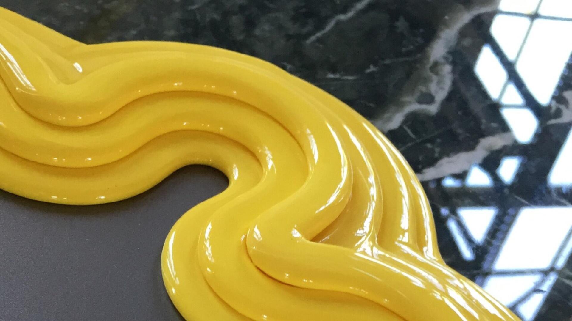 A close-up of a wavy, glossy yellow frame on the corner of a mirror, with a dark marble surface in the background.