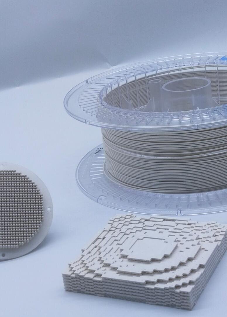 Eine Spule mit weißem 3D-Druckerfilament liegt neben zwei 3D-gedruckten Objekten: einem runden Gitter mit gleichmäßig verteilten Stiften und einem quadratischen Stück mit einem geschichteten, gestuften geometrischen Design.