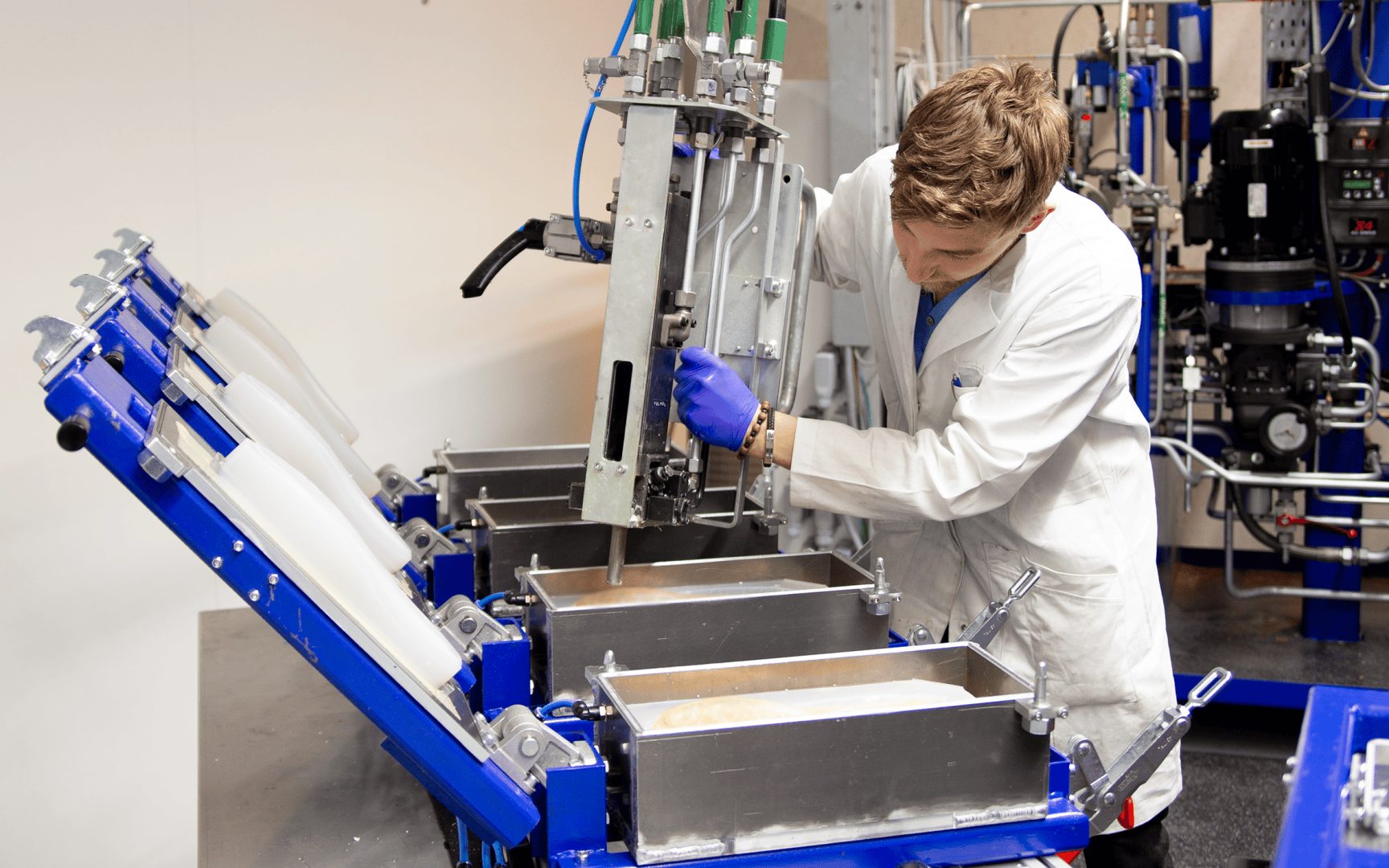 Eine Person in einem weißen Laborkittel und blauen Handschuhen bedient in einem Industrielabor eine Maschine, die Flüssigkeit in rechteckige Metallformen gießt.