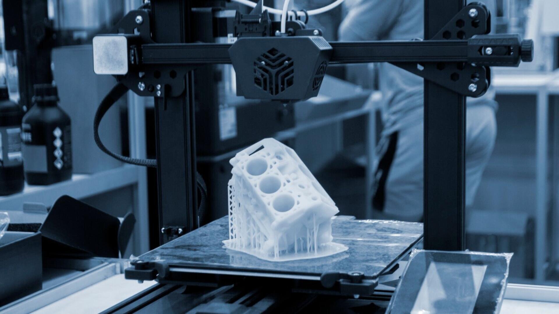 A 3D printer is printing a detailed, white model of an engine block in a workshop setting. The printer head is in motion and the workspace contains various equipment in the background.