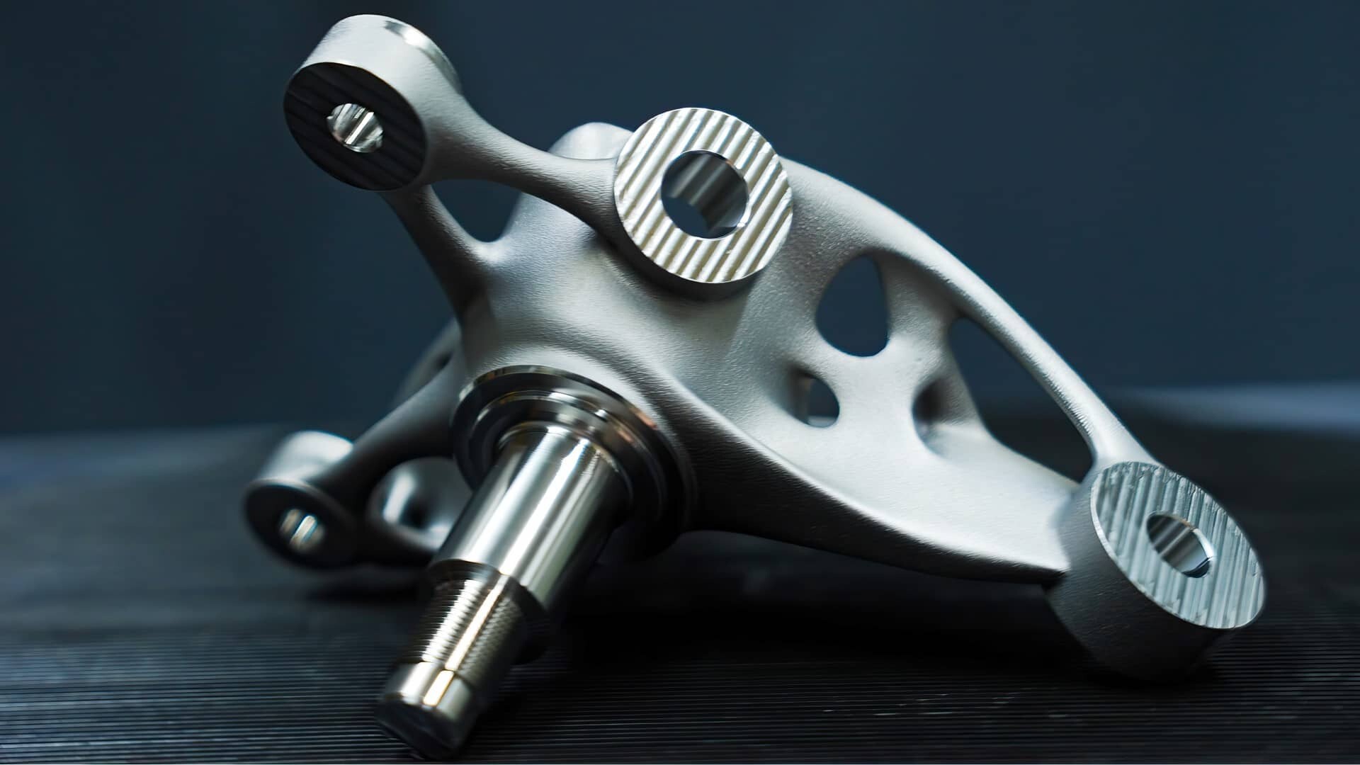 A close-up of a metallic, intricately designed mechanical part with smooth and textured surfaces, featuring circular holes and a central shaft, resting on a dark surface against a dark background.
