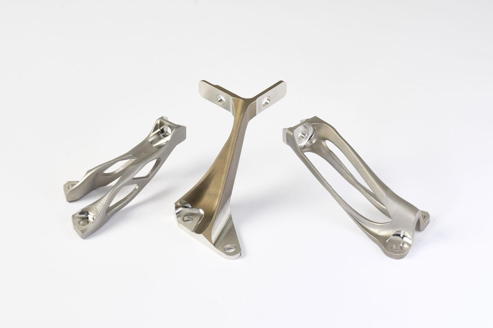 Drei metallische, 3D-gedruckte Strukturhalterungen mit komplizierten, geometrischen Mustern werden auf einer weißen Oberfläche präsentiert. Jede Halterung hat eine einzigartige Form mit sanften Rundungen und offenen, gitterartigen Abschnitten.