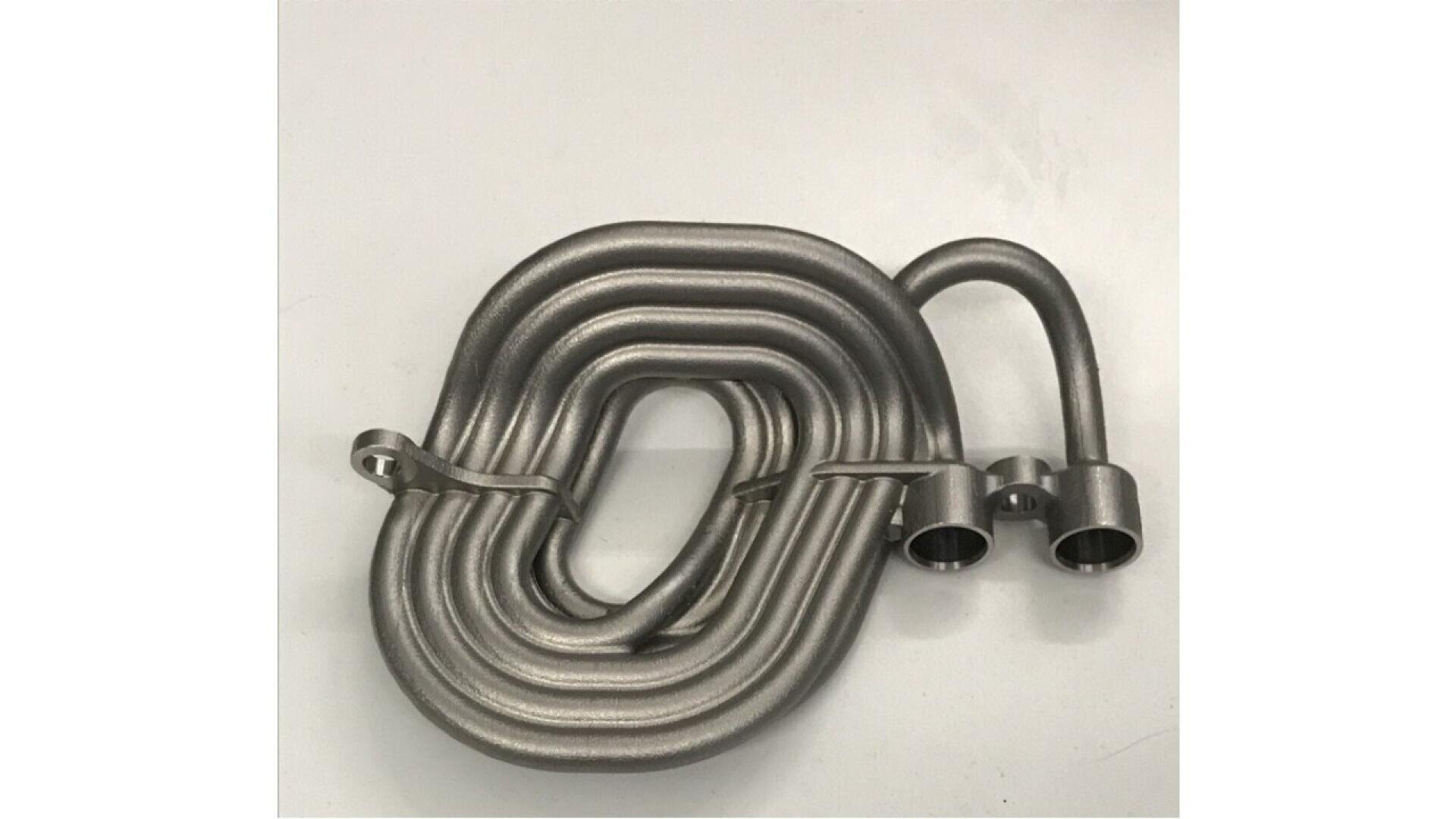 A coiled metallic tube with two open ends and a small bracket attached, placed on a plain white surface. The tube is tightly wound in an oval shape.
