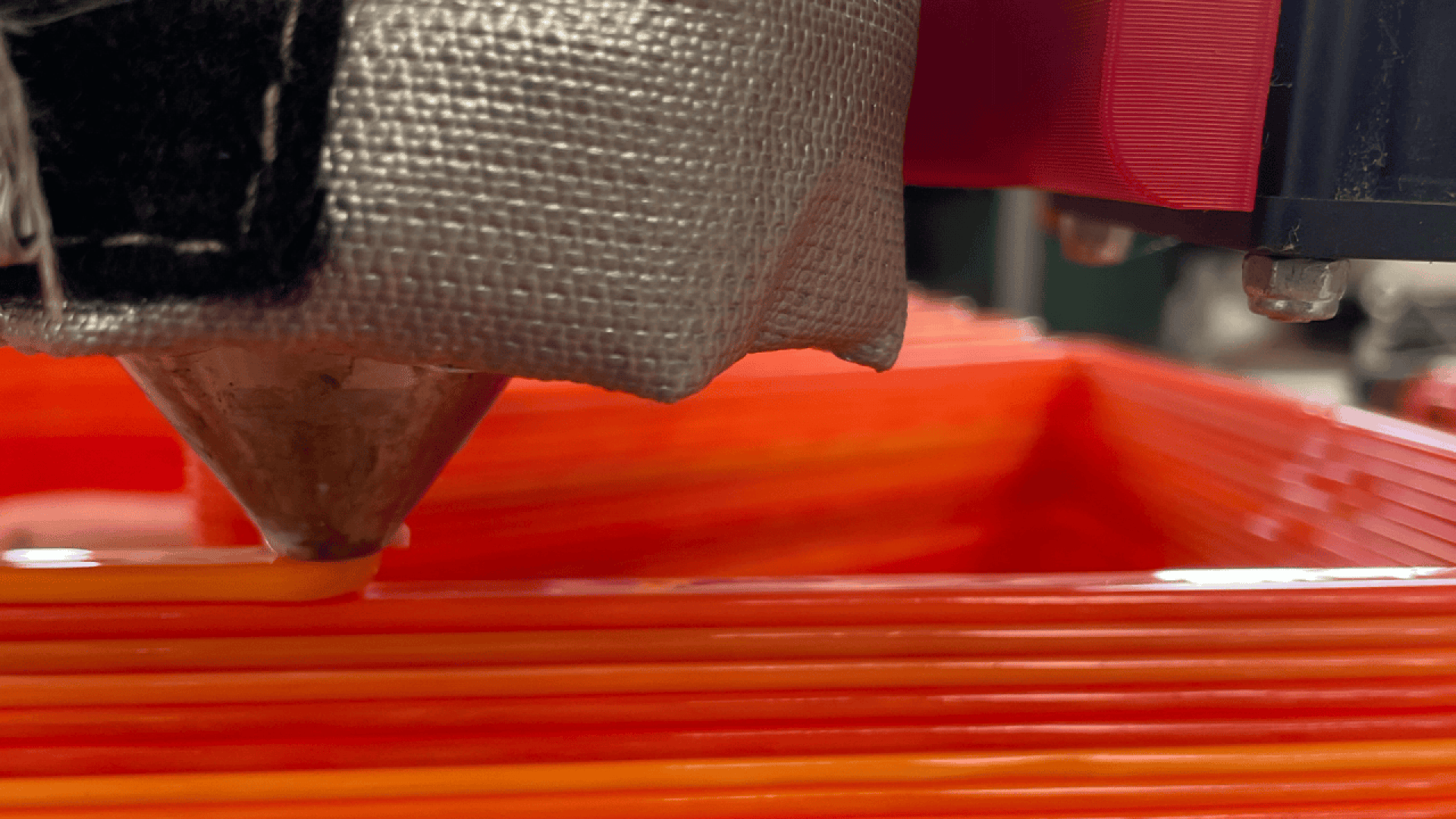 A close-up of a 3D printer nozzle extruding orange filament, creating layers to form a rectangular object. The printhead and part of the machine are visible in the foreground.