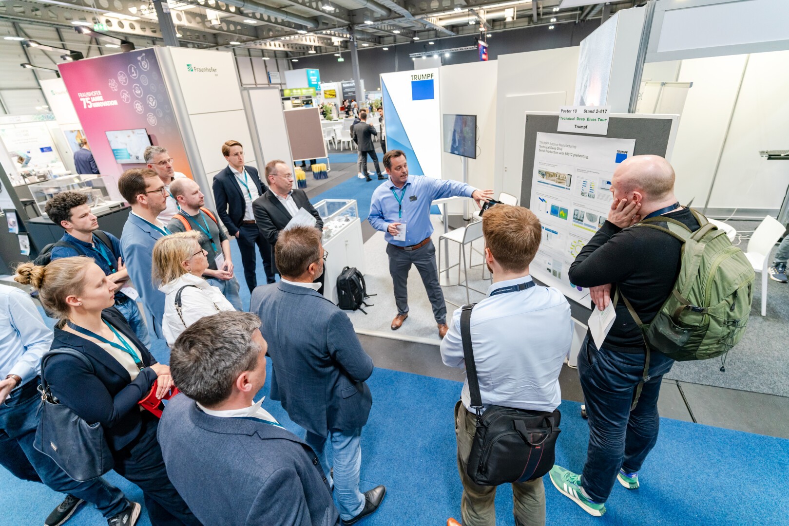 A group of people stands around a presenter who is explaining information displayed on a large poster at a conference or trade show. Attendees listen attentively in a well-lit, modern exhibition hall.