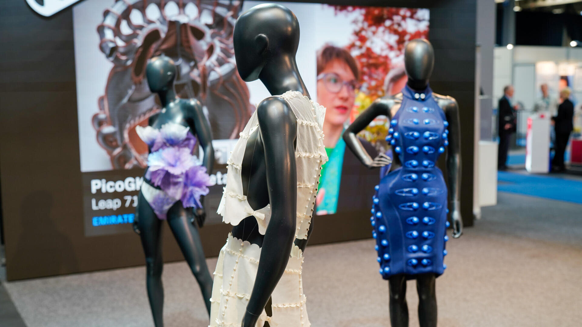 Drei Schaufensterpuppen präsentieren futuristische, strukturierte Outfits in einem modernen Ausstellungsraum; eine trägt ein weißes Rüschenset, eine andere ein lila Blumenmuster und die dritte ein blaues Kleid mit erhabenen Details. Im Hintergrund ist ein Bildschirm mit einer Frau zu sehen.