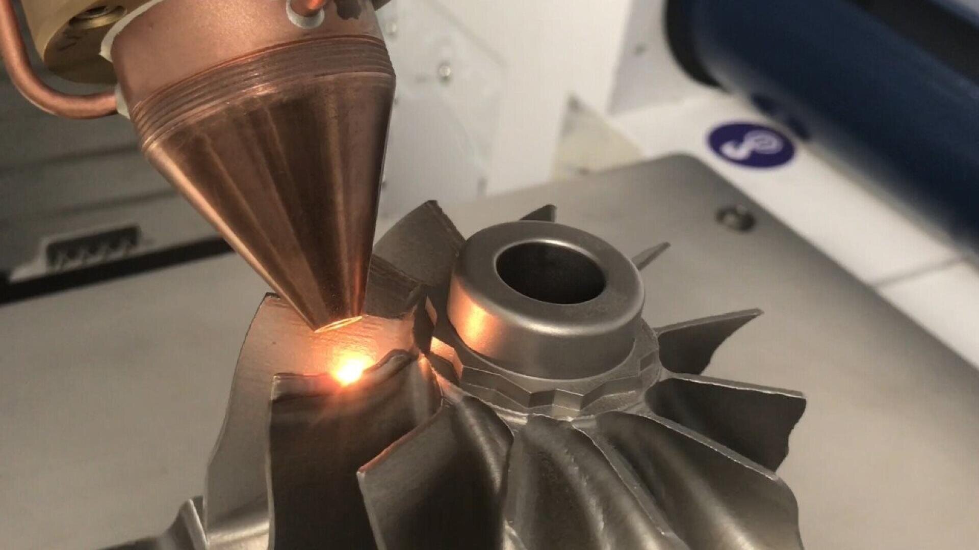 A laser machine repairs a metal turbine blade by directing a focused beam onto its surface, highlighting the area being worked on with a bright orange glow.
