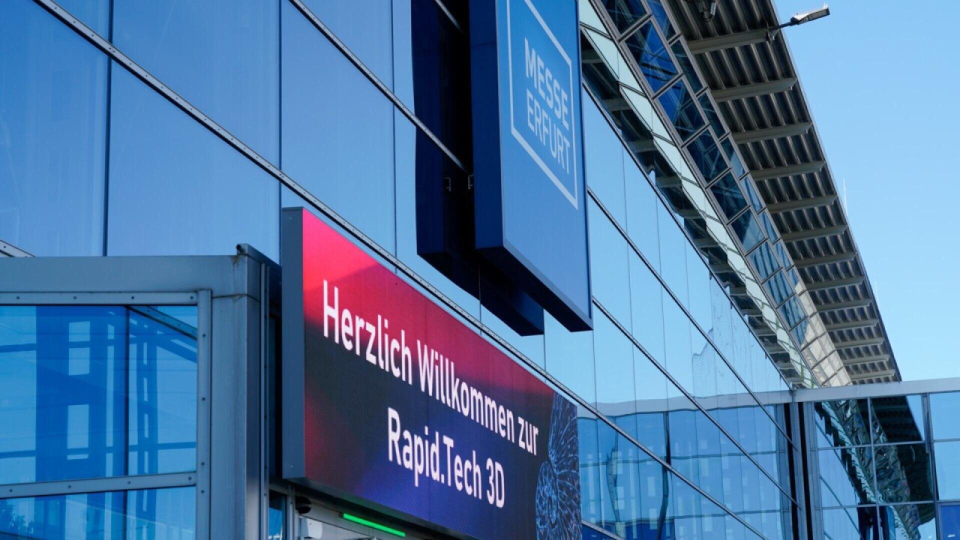 A digital sign at Messe Erfurt displays Herzlich Willkommen zur Rapid.Tech 3D in German, welcoming visitors to the event. The modern glass building reflects sunlight.