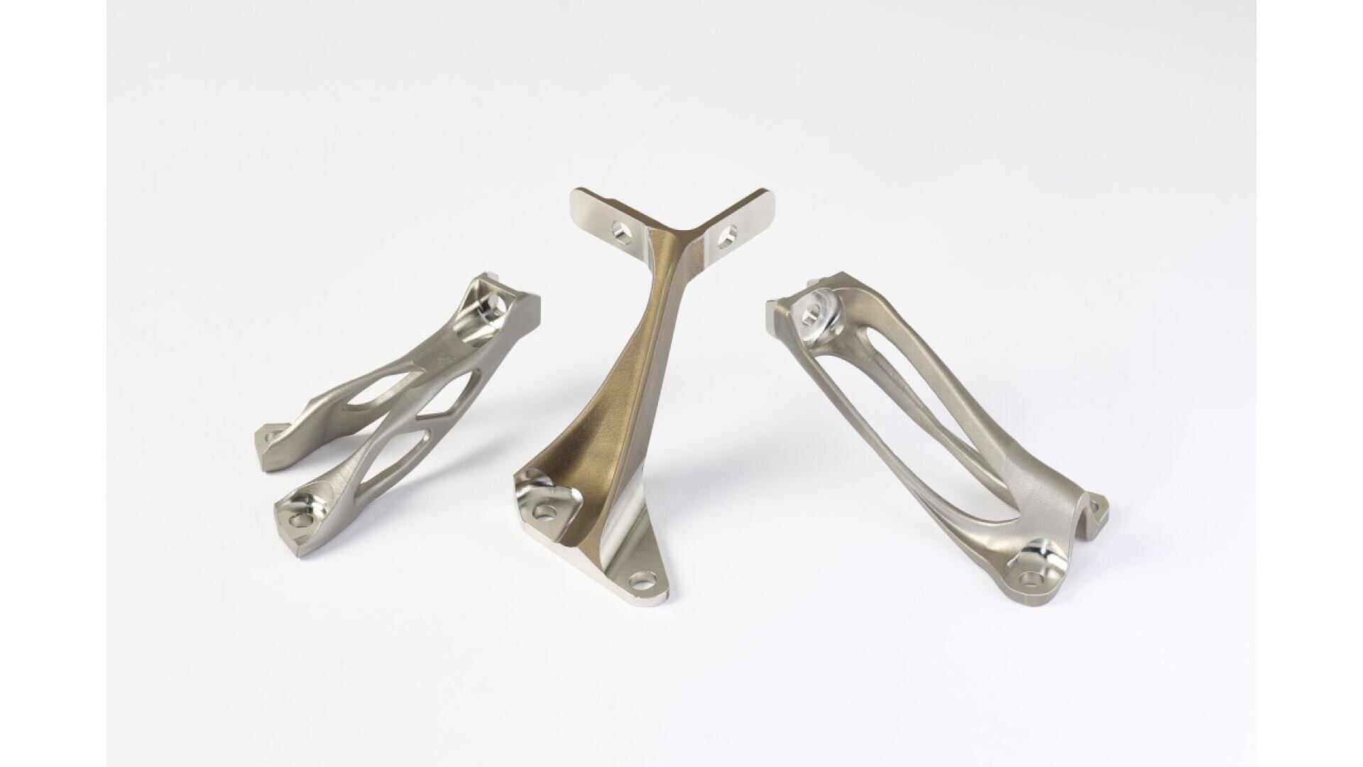 Three metallic, geometrically complex mechanical parts are displayed on a white background. The parts feature smooth, contoured surfaces and open lattice-like structures, suggesting they are lightweight, possibly 3D-printed components.