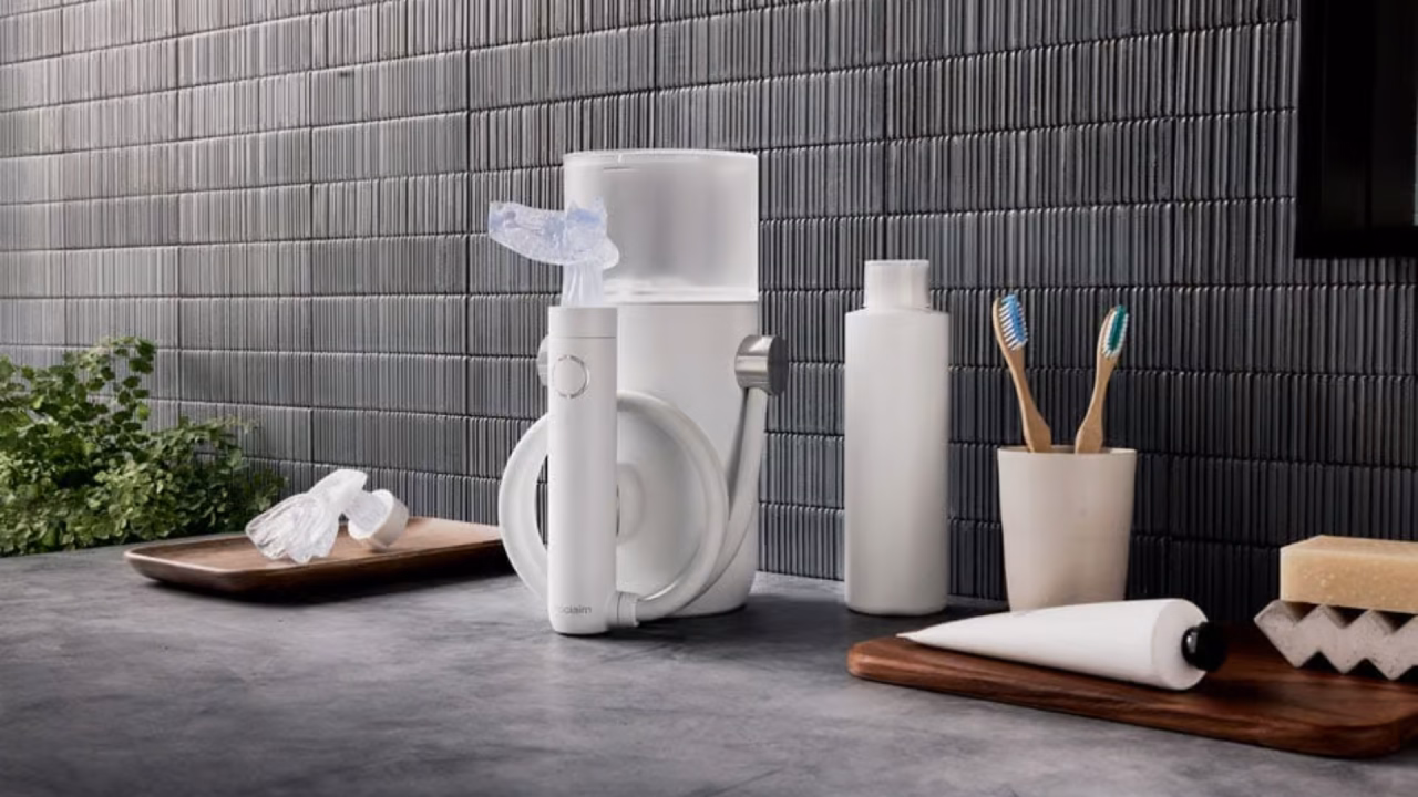 A modern bathroom countertop with an electric water flosser, a water bottle, two toothbrushes in a cup, a towel, soap, and a tray, set against a textured gray tile wall.