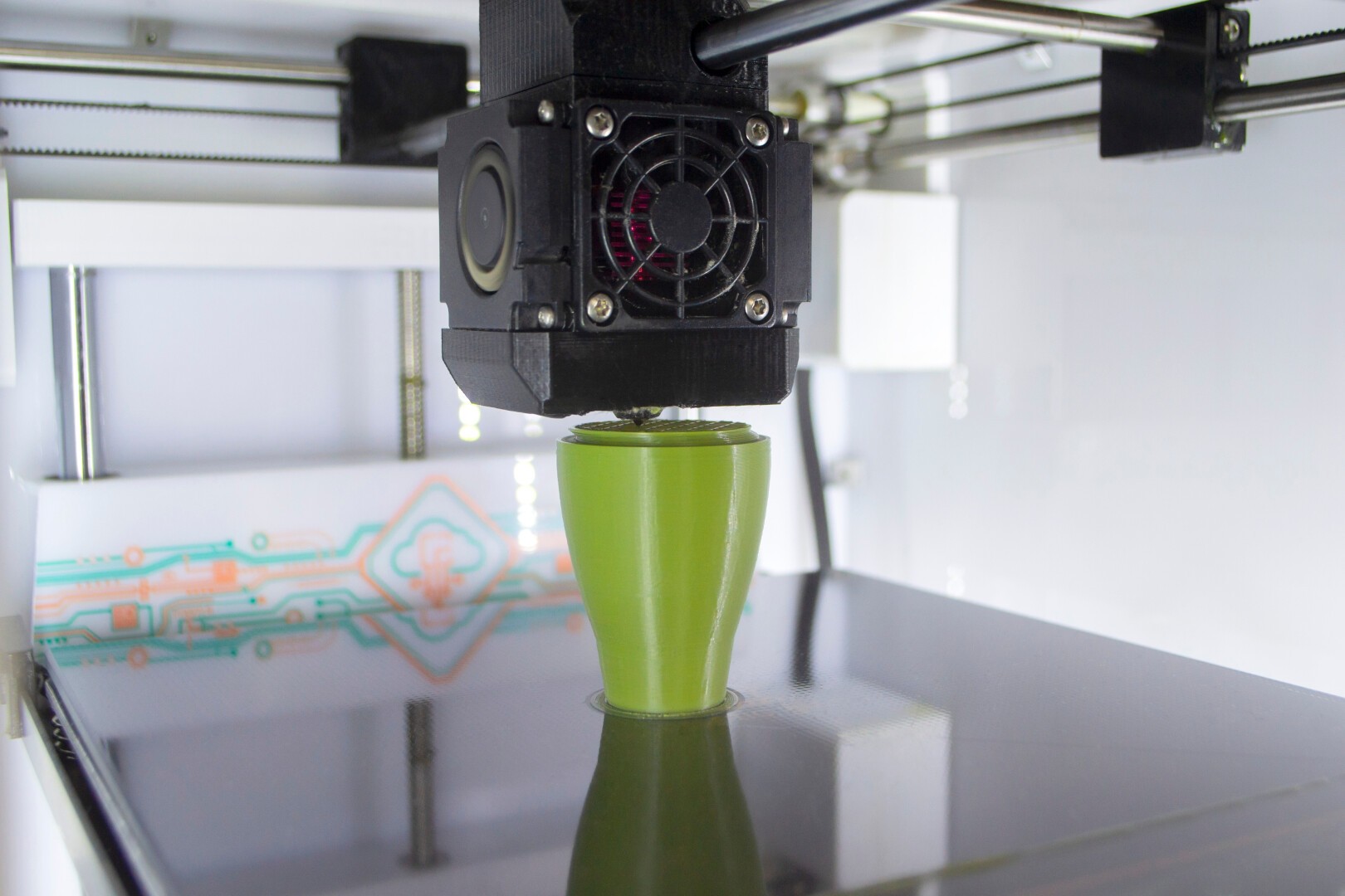 A 3D printer is creating a small green vase-like object on its platform, with the print head in motion and visible electronic components in the background.