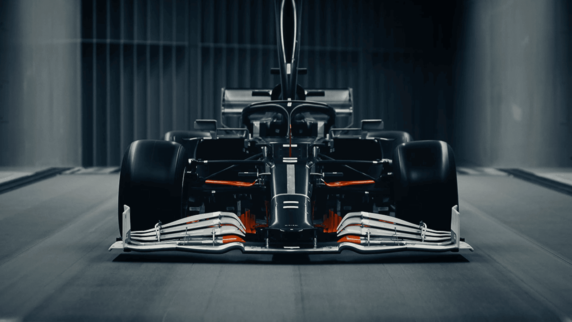 Ein schnittiger, moderner Formel-1-Wagen, der in einem schwach beleuchteten Windkanal von vorne betrachtet wird und seinen aerodynamischen Frontflügel, die breiten Reifen und die futuristischen Designdetails mit roten und schwarzen Akzenten zur Schau stellt.