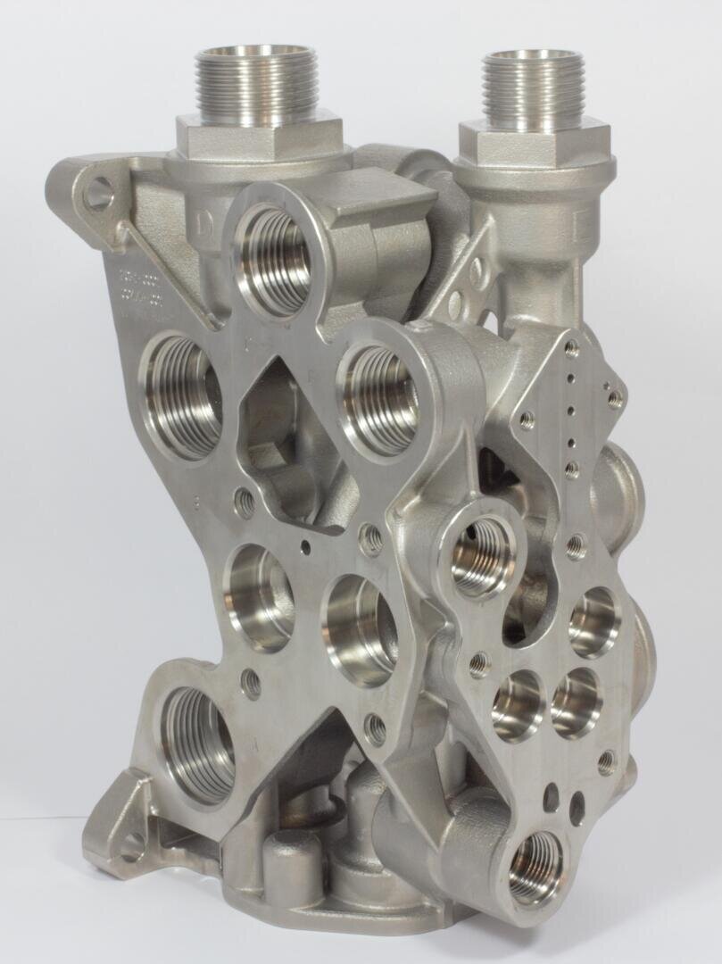 Ein präzisionsgefertigtes Metallbauteil mit mehreren Gewindeanschlüssen und Löchern, wahrscheinlich Teil eines hydraulischen oder mechanischen Systems, das aufrecht vor einem schlichten weißen Hintergrund steht.
