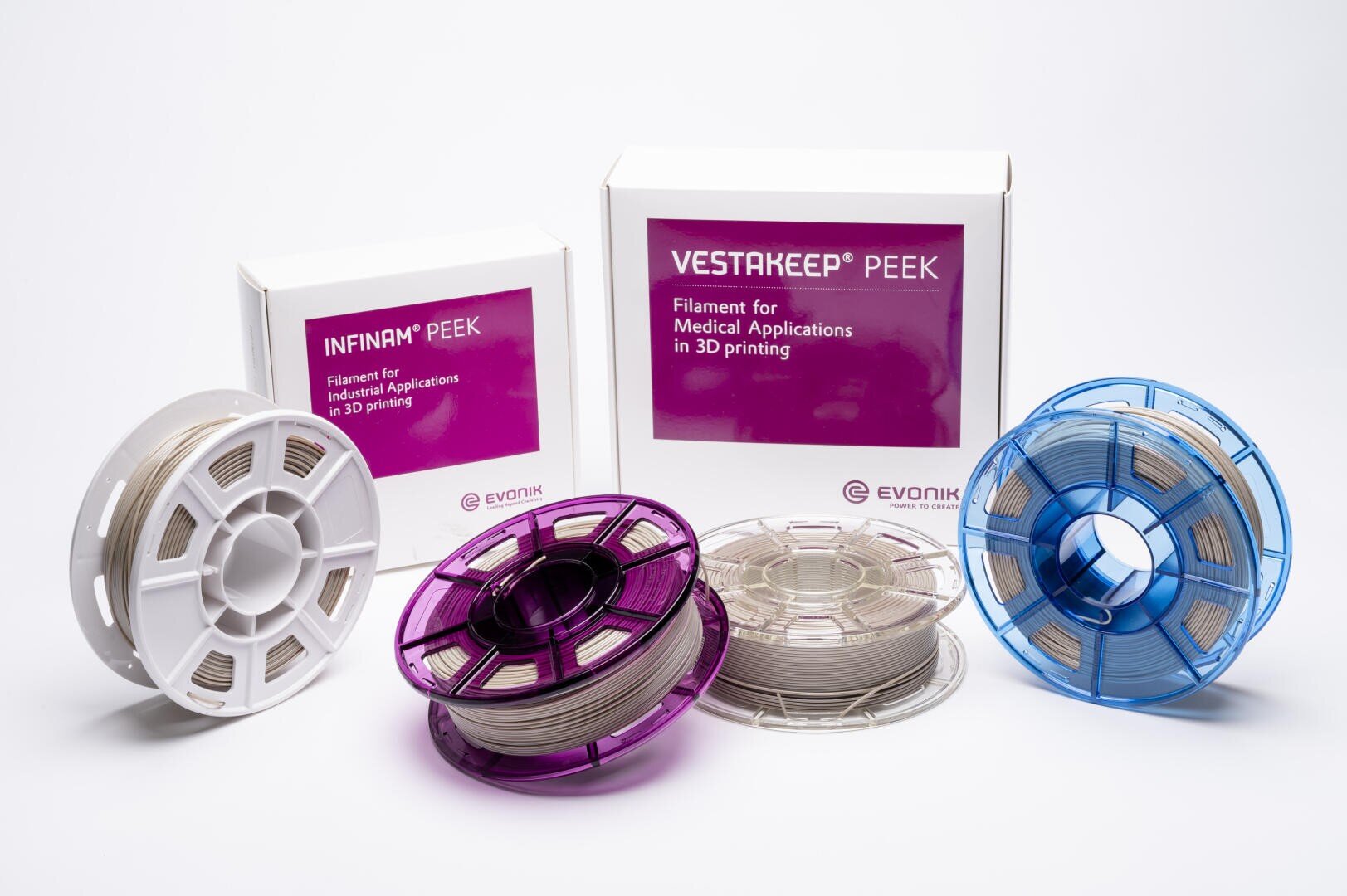 Drei Spulen 3D-Druckfilament (weiß, lila, blau) stehen vor zwei Kartons mit der Aufschrift INFINAM PEEK und VESTAKEEP PEEK, Filamente für den industriellen und medizinischen 3D-Druck von Evonik.