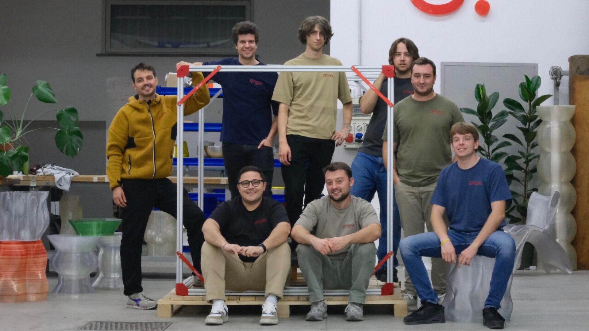 Eight young adults pose together in a workshop, some standing and some sitting on a wooden pallet around a cube frame. They are smiling and casually dressed; various tools, plants, and 3D-printed objects are visible in the background.