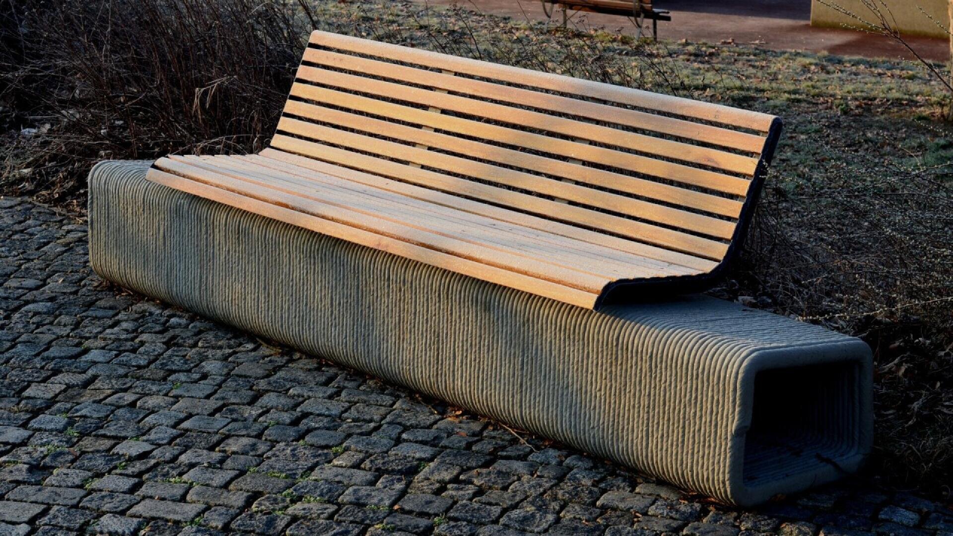 A modern outdoor bench with a wooden slatted seat and a sturdy, rectangular concrete base sits on a cobblestone surface near dried plants.