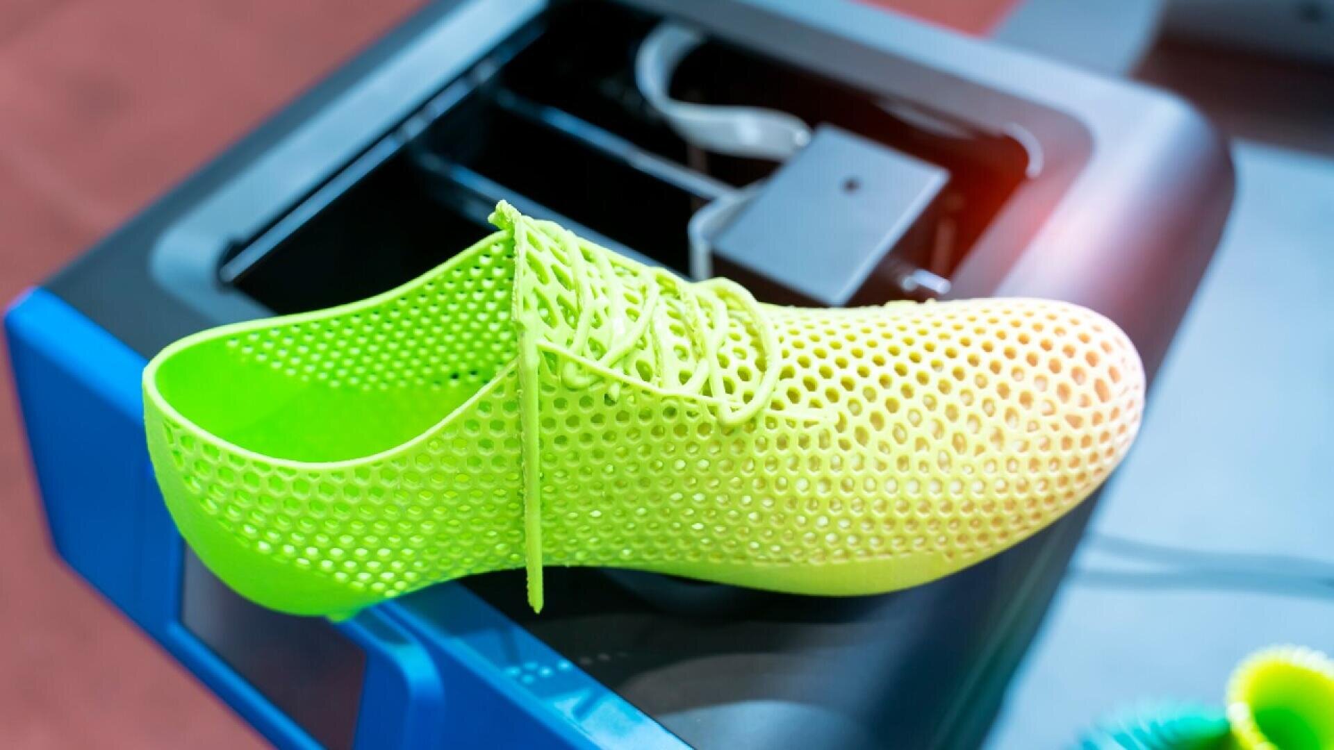 A neon green, 3D-printed mesh shoe with laces sits on a 3D printer platform, showcasing its lightweight, breathable design.