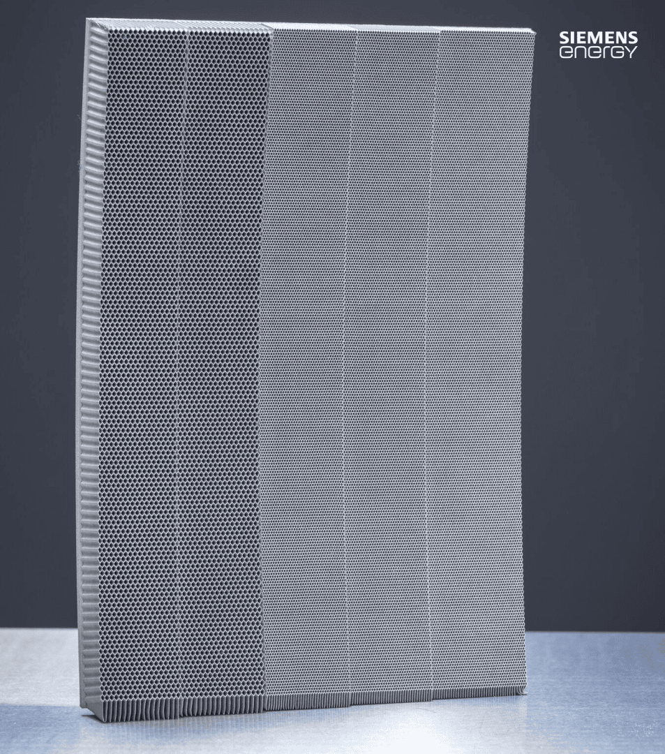 A metallic, rectangular filter with a grid-like pattern stands upright on a metallic surface against a plain background. The Siemens Energy logo appears in the upper right corner.