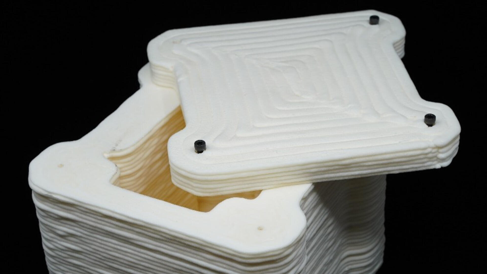 A white, square-shaped 3D-printed container with a textured, layered design sits with its matching lid slightly ajar. Four black screws are visible at the lids corners, and the background is black.