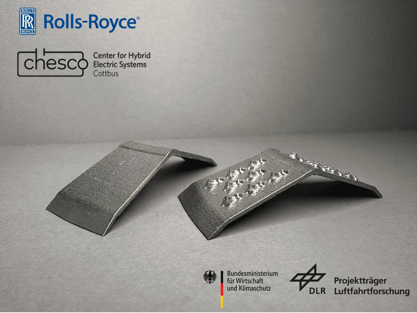 Two metallic components are displayed side by side on a gray surface; the left piece is smooth, while the right has a rough, uneven top. Various company and organization logos are visible in the image’s upper and lower areas.