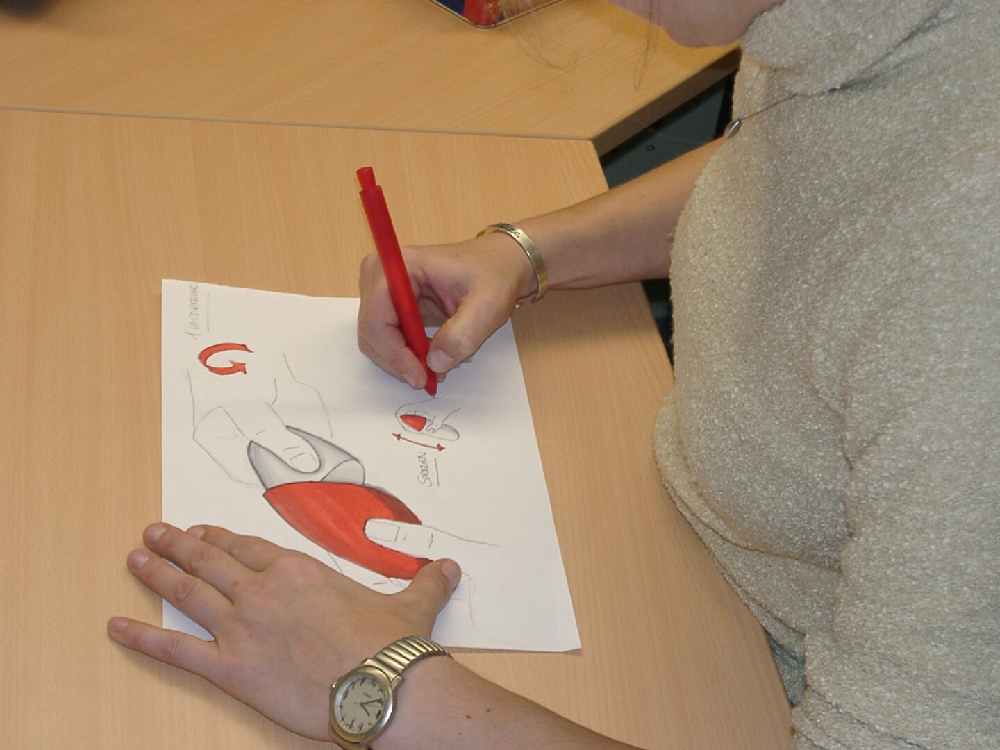 Eine Person mit einem beigen Pullover und einer Armbanduhr sitzt an einem Schreibtisch und malt mit einem roten Stift ein Diagramm der Anatomie einer Hand auf ein Blatt Papier.