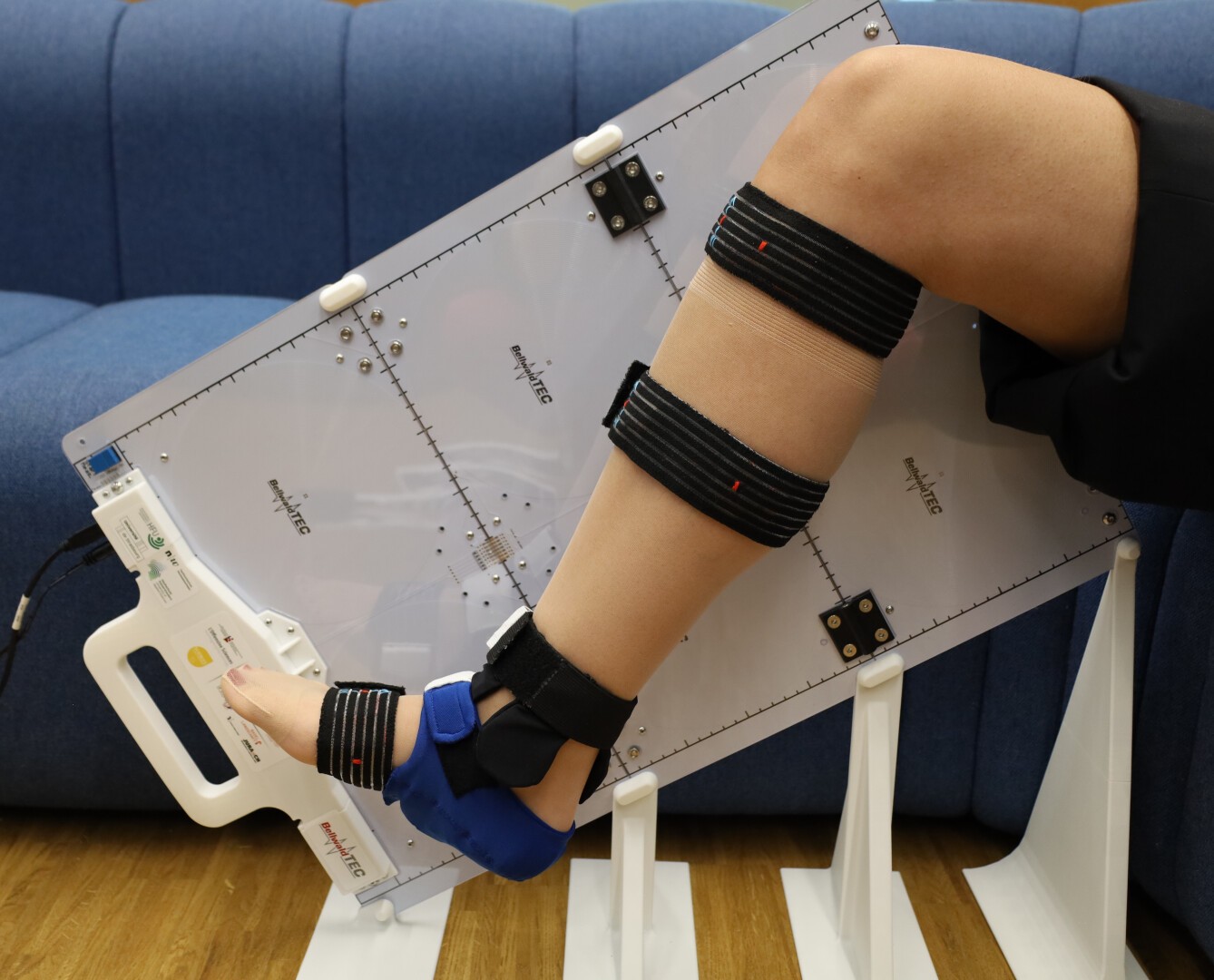 A person’s leg, fitted with black elastic straps and a blue brace, is positioned against a transparent medical device, likely for physical therapy, in front of a blue couch.