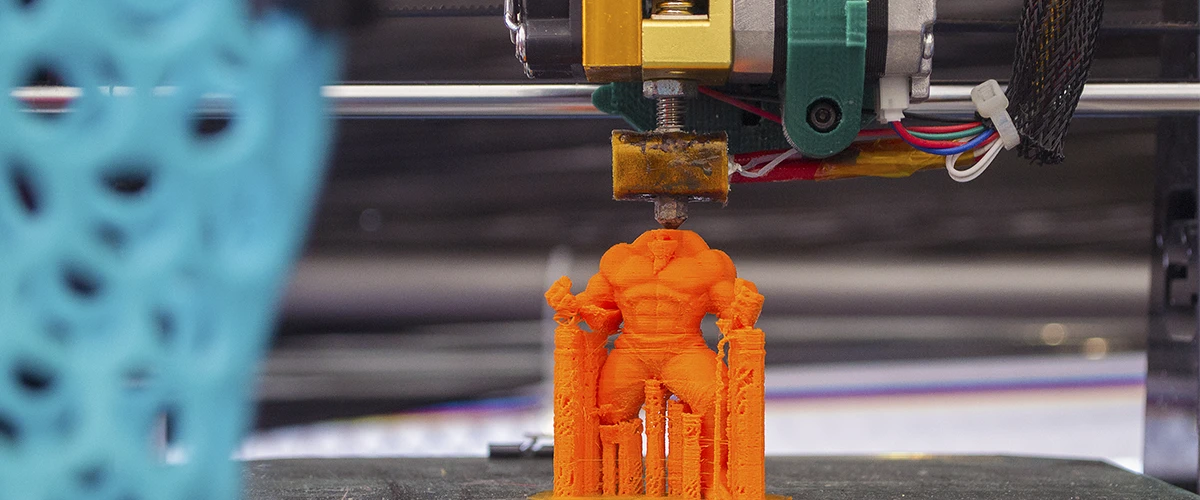 A 3D printer creates a detailed orange model depicting a muscular human figure. The print head is in action and the fine filament layers of the object are clearly visible. In the background, other elements of the printing process can be seen out of focus, underlining the technical focus.