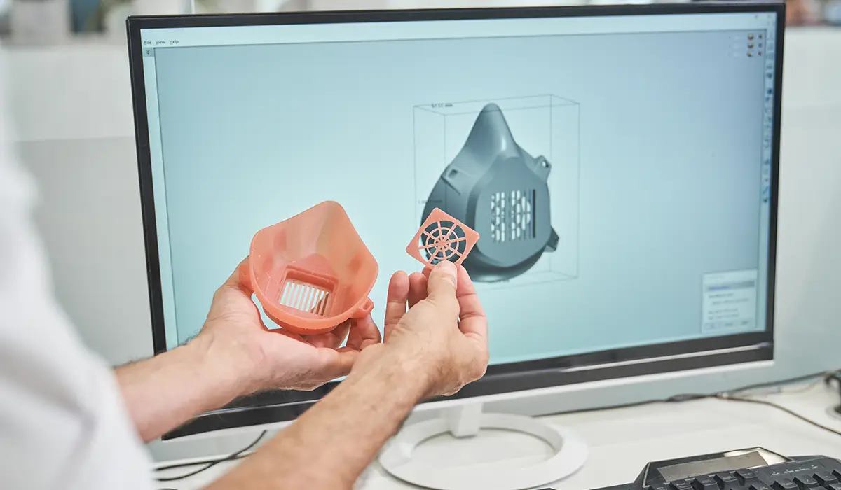 A person holds two 3D-printed components of a respirator mask and compares them with the digital 3D model shown on the monitor in the background.