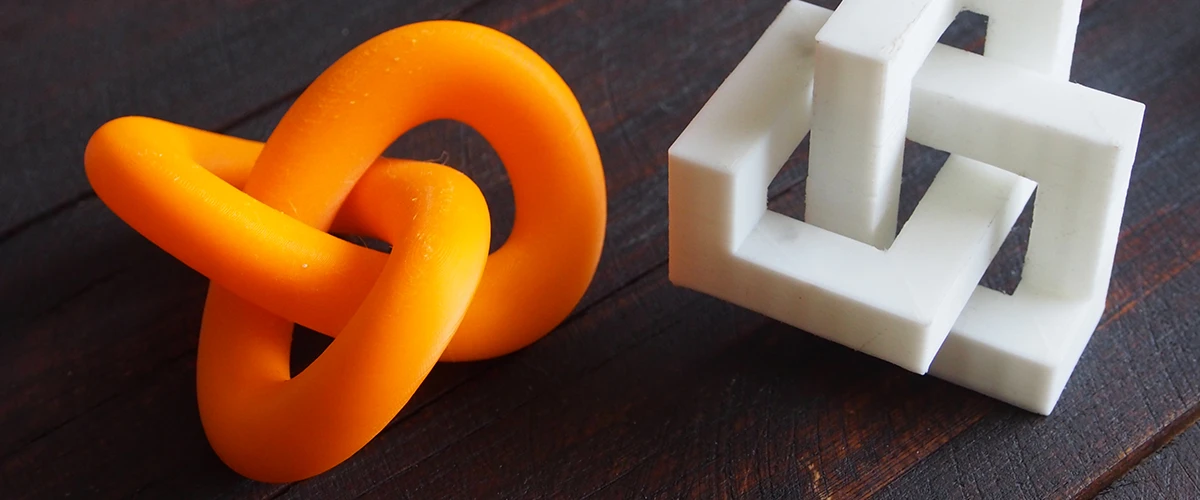 Two 3D printed objects lie on a dark wooden surface: an orange, intricate knot pattern and a white, geometric structure with nested squares. Both models demonstrate the precision and versatility of 3D printing in creating complex shapes.
