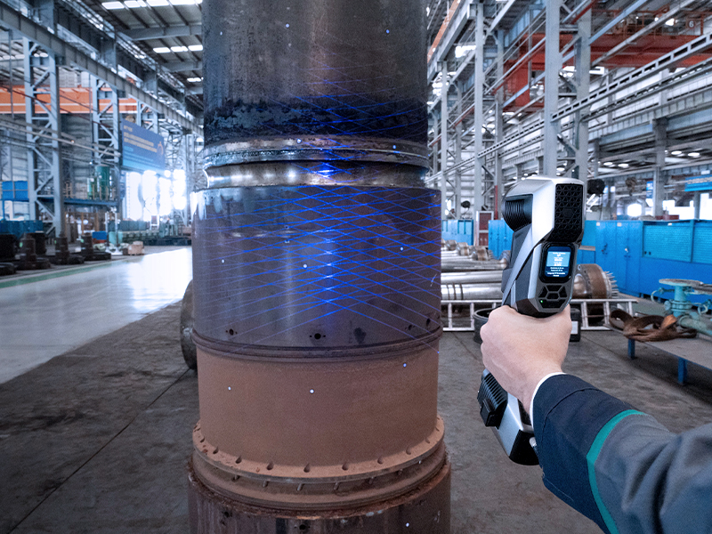 A person uses a handheld 3D scanner emitting blue grid lines to scan a large cylindrical metal object inside an industrial factory.