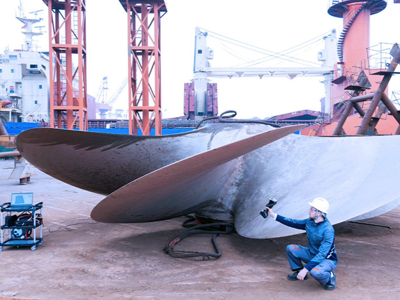 A worker in a hard hat crouches beside a large ship propeller, inspecting it with a handheld device at a shipyard with cranes and industrial equipment in the background.