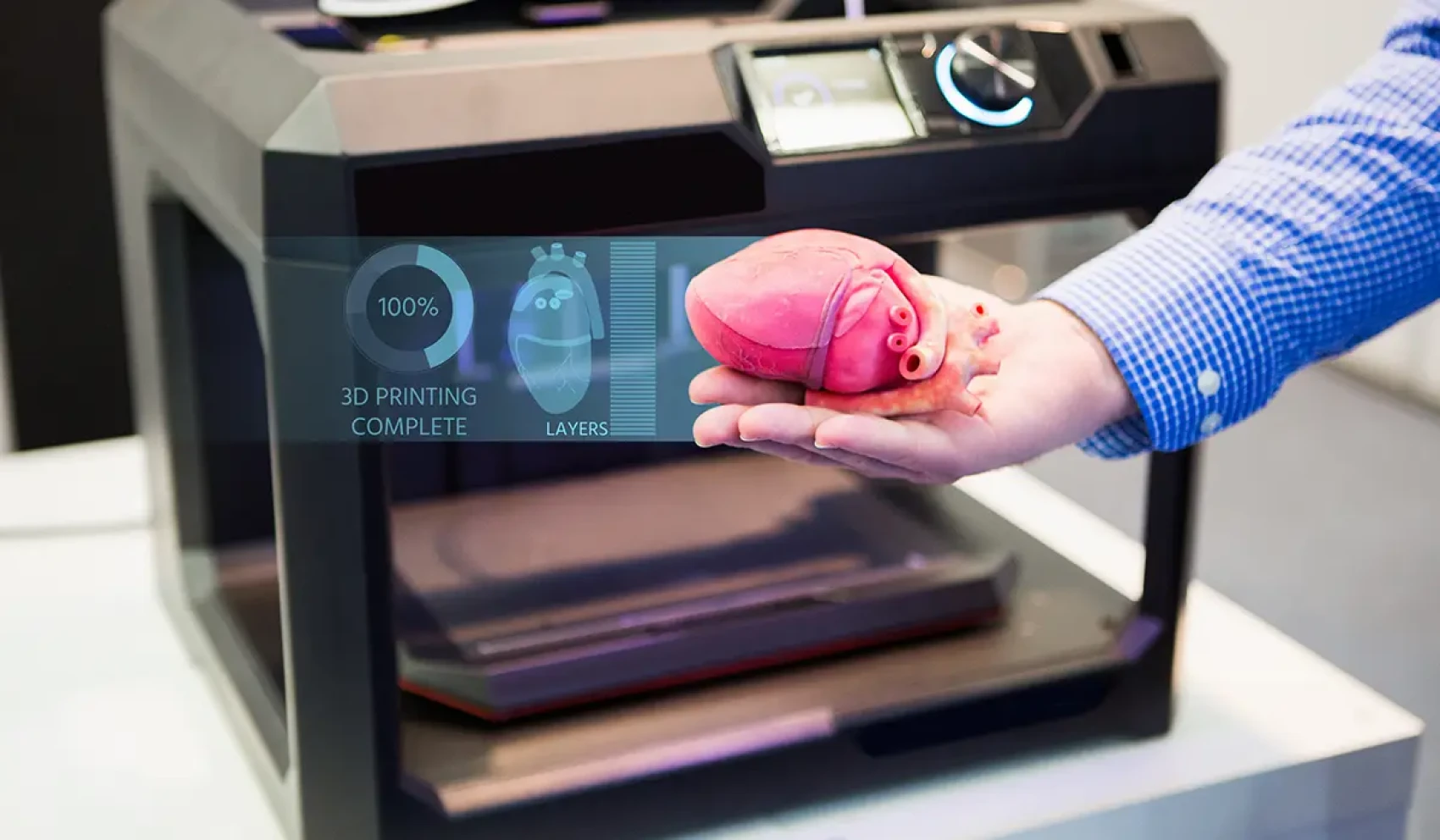 A person holds an artificial heart model that appears to have been freshly 3D-printed. A digital display shows the completed printing process at 100% progress.