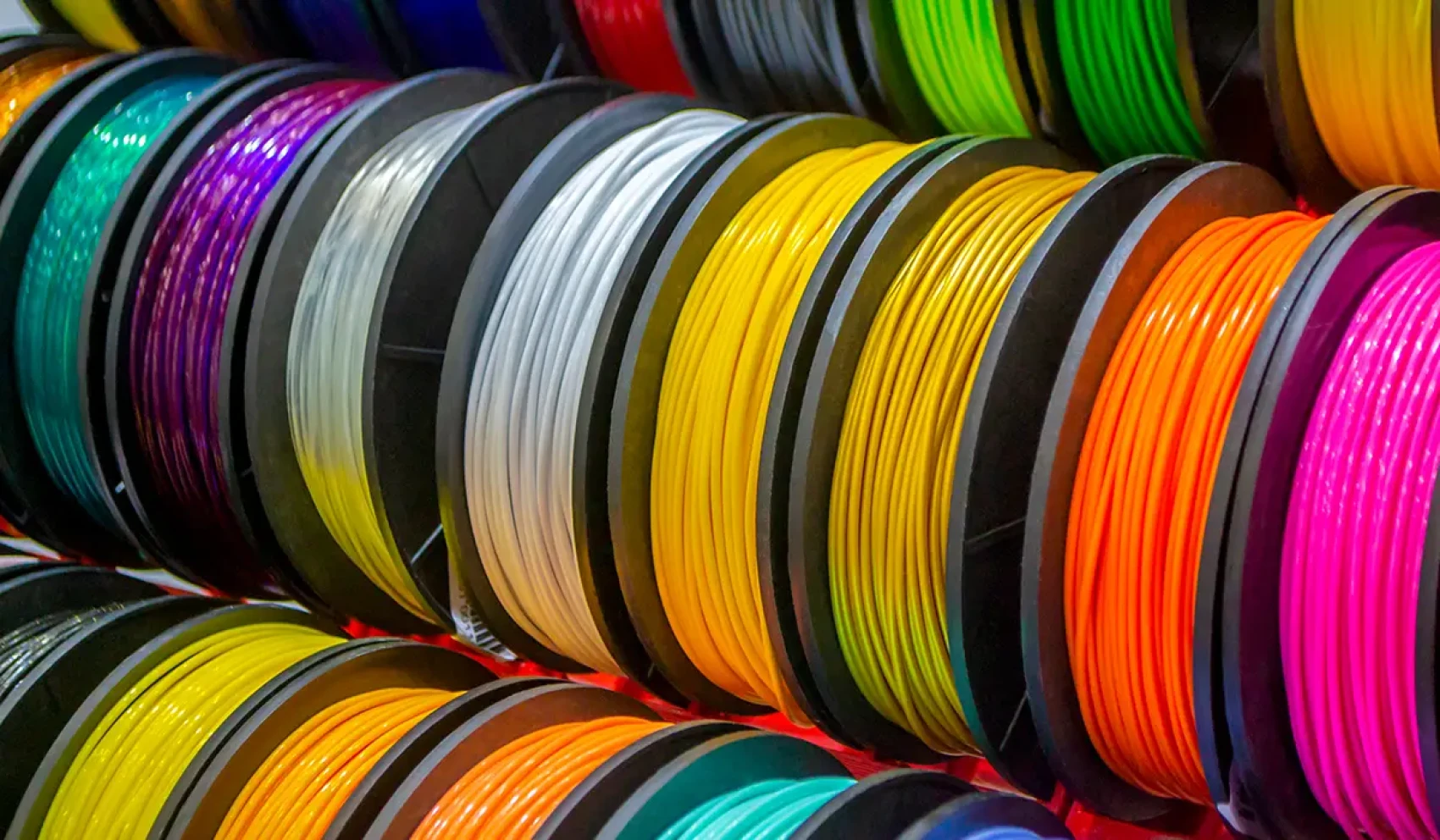 Many colorful filament spools in various colors are neatly lined up next to each other. The scene showcases a wide selection of materials for 3D printing.