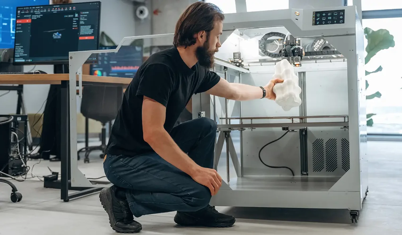 A man in black clothing removes a large, white 3D-printed object from an industrial 3D printer. In the background are several screens with technical software.