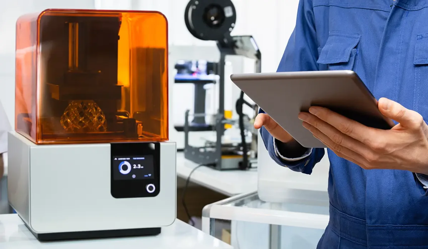 A modern 3D printer with an orange cover stands on a table, next to it, a person in blue work clothes holds a tablet. Other technical devices can be seen in the background.